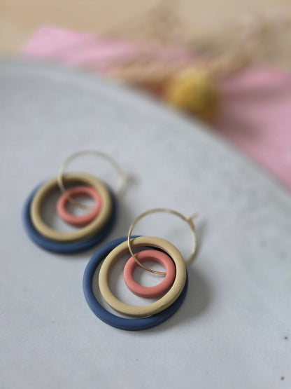 Drie ringen in verschillende groottes van diep blauw, terracotta en mosterd geel oorbellen aan een gouden hoop.
